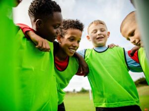 vier jongens met groene hesjes op een sportveld in een kring met armen over elkaar geslagen